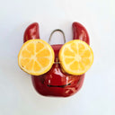 Ceramic sculpture of a red devil head, with a smooth cute face and softly curved horns. On its eyes are slices of lemons.