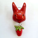 Glazed ceramic sculpture of a red devil head, cute and simplistic in form with long horns. From it hangs a small strawberry.
