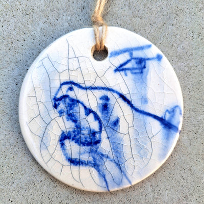 Round ceramic pendant with a cracked glaze finish and a blue watercolor style illustration of Godzilla crying out, wiht a small helicopter overhead.