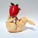 Glazed ceramic sculpture of a white bunny, laying on its back with a large red strawberry balanced on its feet.