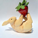 Glazed ceramic sculpture of a white bunny, laying on its back with a large red strawberry balanced on its feet.