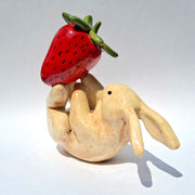 Glazed ceramic sculpture of a white bunny, laying on its back with a large red strawberry balanced on its feet.