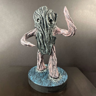 Textured sculpture in muted tones of a character appearing to be a driftwood creature, with no mouth and only eyes. It stands on a base and has its arms extended up or out.