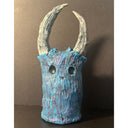 Textured clay sculpture of a blue head made to look like a wooden tree trunk with no features aside from hollowed eyes and nose holes. Large asymmetrical horns grow atop.