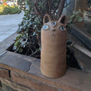 Ceramic cat-shaped totem of sorts with 3 blue eyes on a ledge with plants in the background