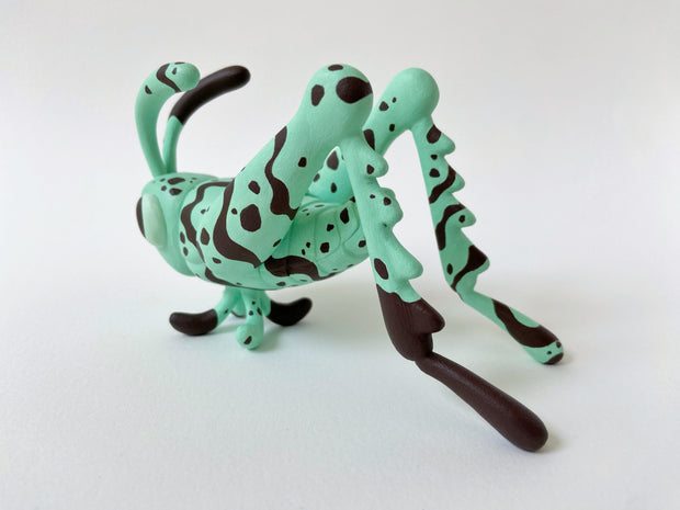 Sculpture of a mint green colored grasshopper with large cute cartoonish eyes and chocolate colored stripes and dots all over its body.