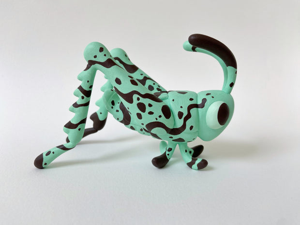 Sculpture of a mint green colored grasshopper with large cute cartoonish eyes and chocolate colored stripes and dots all over its body.