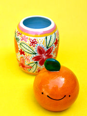Ceramic sculpture comprised of 2 separate parts. Top is a rounded orange with a leaf and smiling cartoon face. Base is a rounded vase with many colorful paintings of fruits and flowers.