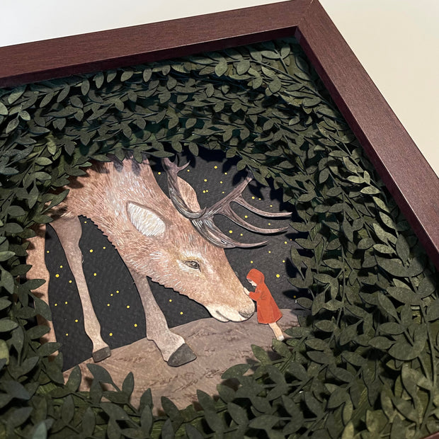 Layered cut paper diorama style sculpture in an open wooden frame, with many layers of leaves revealing a small scene of a red cloaked child petting the face of a very large Elk, which bows its head to the child.