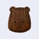 A wooden capybara head shaped bottle opener, flat with the design taking up the whole of the surface, featuring a dark brown wood grain texture, black "stitched" border, and simple black facial features, shown on a light gray background.