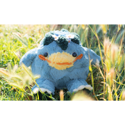 Blue plush doll of a rotund and chubby kappa, a creature with a large smiling yellow beak nose and leaves around a circle atop its head. The plush sits in a grassy field.