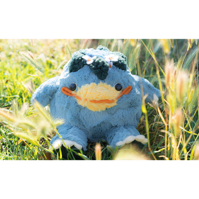 Blue plush doll of a rotund and chubby kappa, a creature with a large smiling yellow beak nose and leaves around a circle atop its head. The plush sits in a grassy field.