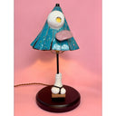 A whimsical table lamp shaped like a stylized triangular head with blue patterning, a pink tongue being stuck out, a white eye, and a white sock and sandal for the base, on a dark round base, set against a solid pink background.