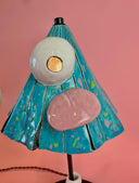 A whimsical table lamp shaped like a stylized triangular head with blue patterning, a pink tongue being stuck out, a white eye, and a white sock and sandal for the base, on a dark round base, set against a solid pink background.
