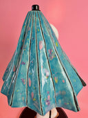 A whimsical table lamp shaped like a stylized triangular head with blue patterning, a pink tongue being stuck out, a white eye, and a white sock and sandal for the base, on a dark round base, set against a solid pink background.