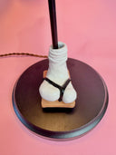 A whimsical table lamp shaped like a stylized triangular head with blue patterning, a pink tongue being stuck out, a white eye, and a white sock and sandal for the base, on a dark round base, set against a solid pink background.