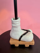 A ceramic novelty figurine shaped like a white sock boot with a black strap, mounted on a tan base and holding a black rod, set against a pink background.