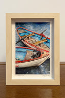 Painting of 2 rowboats, floating atop of sparkling blue water and with the fronts of both touching one another. In the rowboat closest to the viewer sits a black and white cat. Framed.