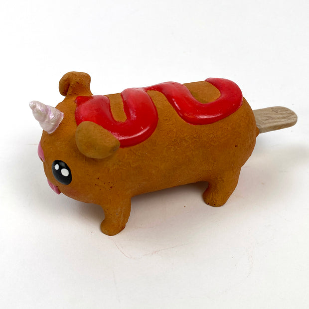 Sculpture of a corn dog, designed to look like a dog with a cute, silly face and a metallic unicorn horn atop its head. It has a squiggle of ketchup on its back.