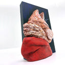 3D sculpture of the profile view of an orange cat, wearing a red hoody. It is attached to a flat dark blue wood panel.