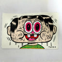 A cartoon-style illustrated face with oversized pink eyes, black hair, a wide open grin, and dripping details on an off-white rectangular background, photographed against a light gray surface.