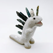 Ceramic sculpture of a white dinosaur with large spikes on its back made out of green and blue stones. A small, cute character stands atop its head and cheers.