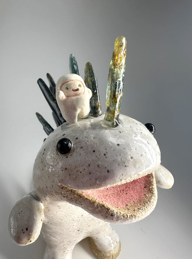 Ceramic sculpture of a white dinosaur with large spikes on its back made out of green and blue stones. A small, cute character stands atop its head and cheers.