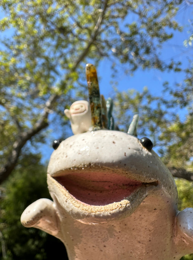 Ceramic sculpture of a white dinosaur with large spikes on its back made out of green and blue stones. A small, cute character stands atop its head and cheers.