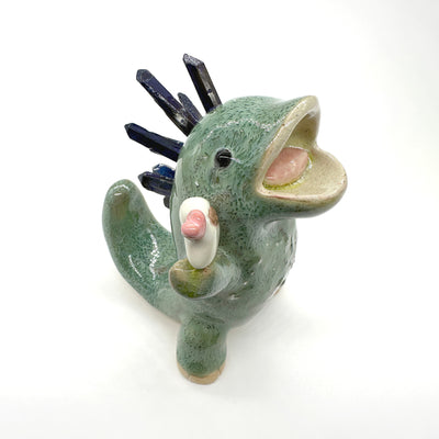 Ceramic sculpture of a green dinosaur with blue stone spikes on its back and an open mouth. It holds a small character in its hand, who holds a heart.