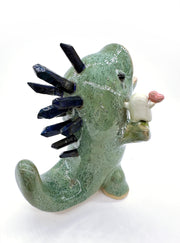 Ceramic sculpture of a green dinosaur with blue stone spikes on its back and an open mouth. It holds a small character in its hand, who holds a heart.