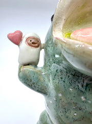 Ceramic sculpture of a green dinosaur with blue stone spikes on its back and an open mouth. It holds a small character in its hand, who holds a heart.