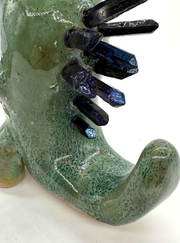 Ceramic sculpture of a green dinosaur with blue stone spikes on its back and an open mouth. It holds a small character in its hand, who holds a heart.