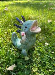 Ceramic sculpture of a green dinosaur with blue stone spikes on its back and an open mouth. It holds a small character in its hand, who holds a heart.