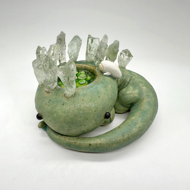 Ceramic sculpture of a green lizard like creature, curled into itself with quarts sticking out of its body. In the center of its head is a pool of water with green plants. A small character looks in.