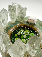 Ceramic sculpture of a green lizard like creature, curled into itself with quarts sticking out of its body. In the center of its head is a pool of water with green plants. A small character looks in.
