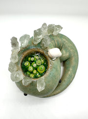 Ceramic sculpture of a green lizard like creature, curled into itself with quarts sticking out of its body. In the center of its head is a pool of water with green plants. A small character looks in.