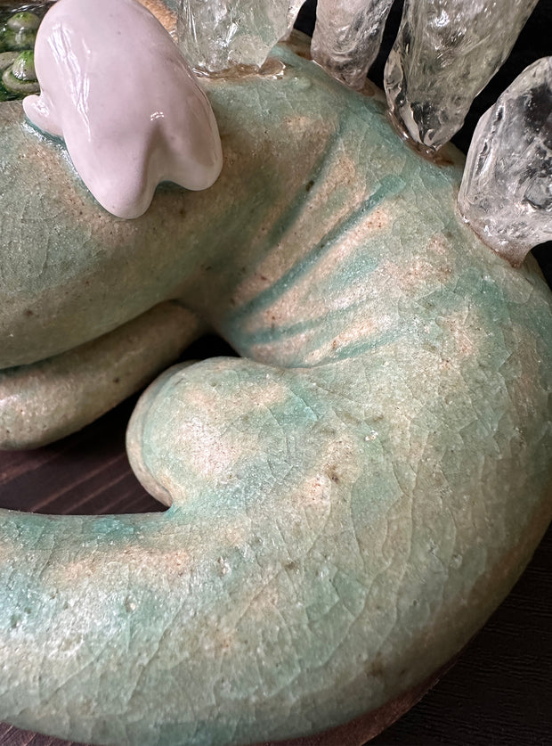 Ceramic sculpture of a green lizard like creature, curled into itself with quarts sticking out of its body. In the center of its head is a pool of water with green plants. A small character looks in.