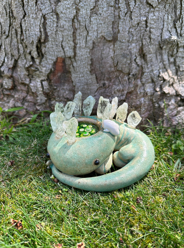 Ceramic sculpture of a green lizard like creature, curled into itself with quarts sticking out of its body. In the center of its head is a pool of water with green plants. A small character looks in.