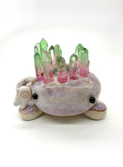 Ceramic sculpture of a brownish purple lizard like creature, curled into itself with quarts sticking out of its body. In the center of its head is a pool of water with green plants. A small character stands on the creature's hand.