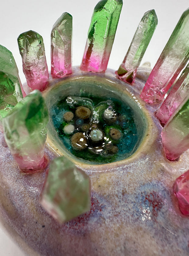 Ceramic sculpture of a brownish purple lizard like creature, curled into itself with quarts sticking out of its body. In the center of its head is a pool of water with green plants. A small character stands on the creature's hand.