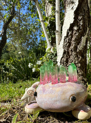 Ceramic sculpture of a brownish purple lizard like creature, curled into itself with quarts sticking out of its body. In the center of its head is a pool of water with green plants. A small character stands on the creature's hand.