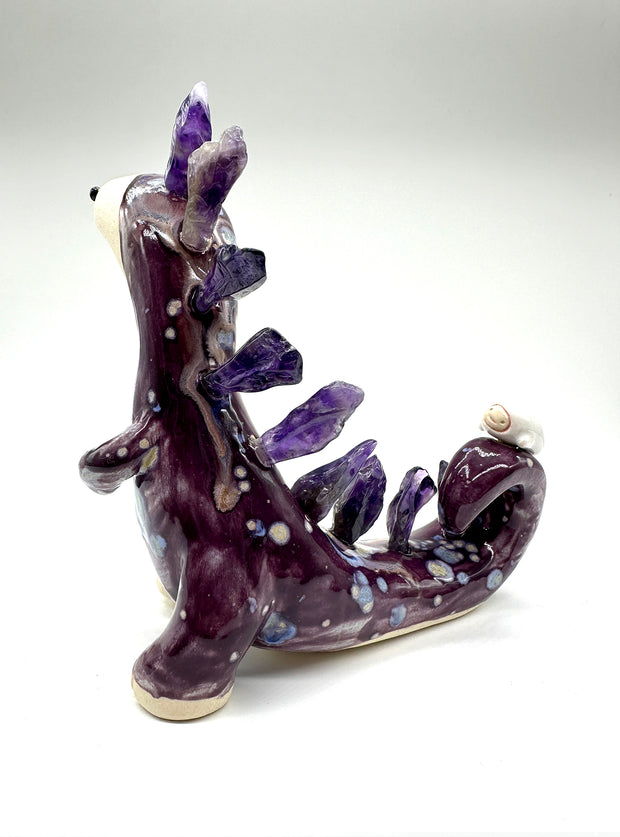 Ceramic sculpture of a lanky purple dinosaur with an off white face. Along its back are spikes made out of amethyst. A small character lays on its tail.