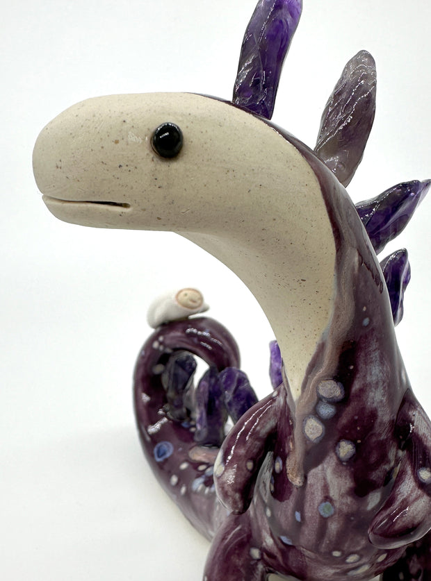 Ceramic sculpture of a lanky purple dinosaur with an off white face. Along its back are spikes made out of amethyst. A small character lays on its tail.