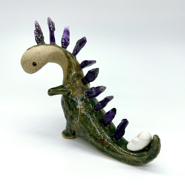 Ceramic sculpture of a lanky green dinosaur with an off white face. Along its back are spikes made out of amethyst. A small character lays on its tail.