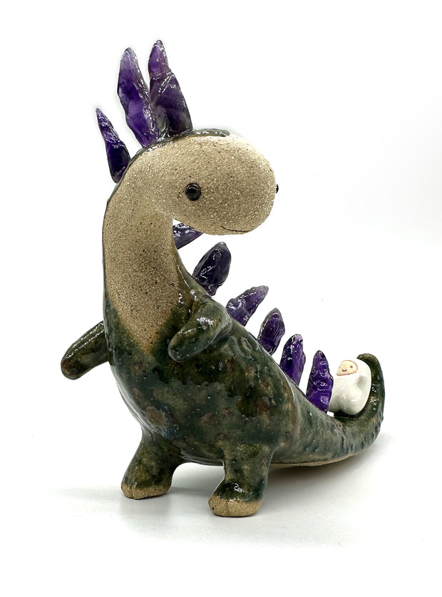 Ceramic sculpture of a lanky green dinosaur with an off white face. Along its back are spikes made out of amethyst. A small character lays on its tail.