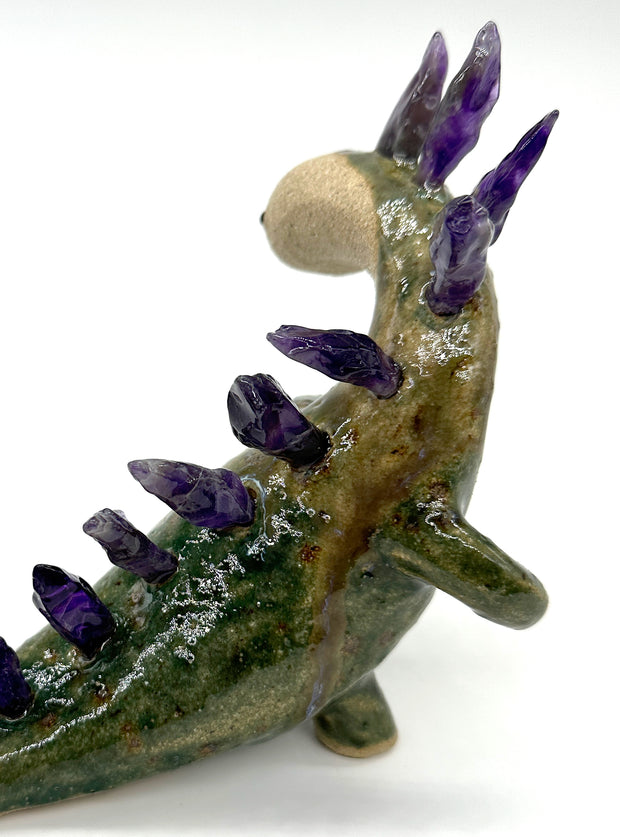 Ceramic sculpture of a lanky green dinosaur with an off white face. Along its back are spikes made out of amethyst. A small character lays on its tail.