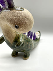 Ceramic sculpture of a lanky green dinosaur with an off white face. Along its back are spikes made out of amethyst. A small character lays on its tail.
