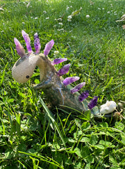 Ceramic sculpture of a lanky green dinosaur with an off white face. Along its back are spikes made out of amethyst. A small character lays on its tail.