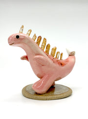 Ceramic sculpture of a pink dinosaur, mid stride, with orange quartz for spikes with a small character clinging to its tail.