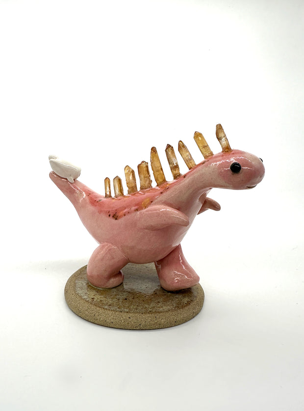 Ceramic sculpture of a pink dinosaur, mid stride, with orange quartz for spikes with a small character clinging to its tail.